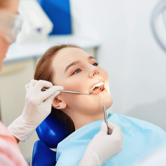 A person reclines in a dental chair while a dental professional wearing gloves uses dental instruments to examine the person’s teeth.