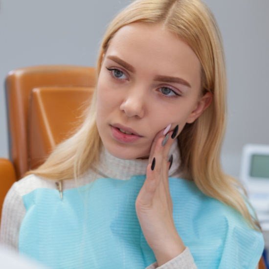 A woman wearing a dental bib sits in a dental chair and gently touches her cheek while looking slightly to the side.