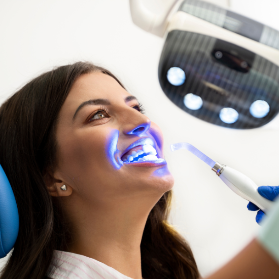 A person reclines in a dental chair with their mouth open while a dental professional uses a handheld device near their teeth under a blue light and an overhead exam lamp.