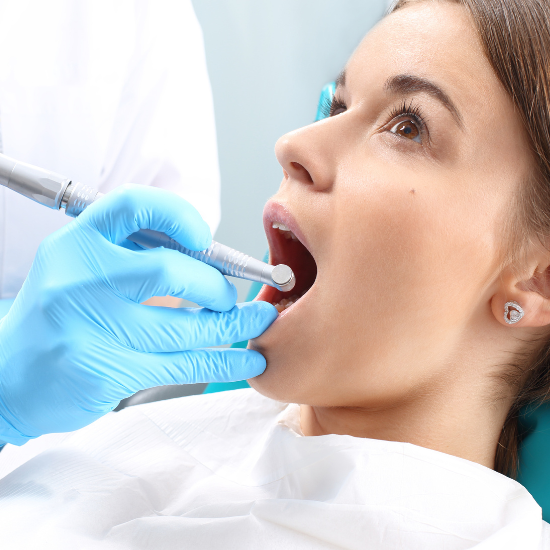 A person reclines in a dental chair while a dental professional wearing blue gloves uses a dental instrument inside the person’s open mouth during an examination or procedure.